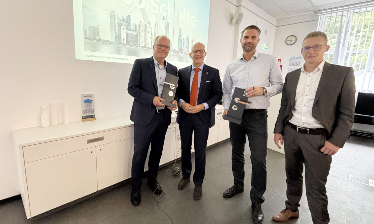 Vier Männer stehen in einem Besprechungsraum vor einer Präsentation mit der Aufschrift „Die Welt von ECO Schulte“. Zwei von ihnen halten schwarze Präsentationsboxen in den Händen.