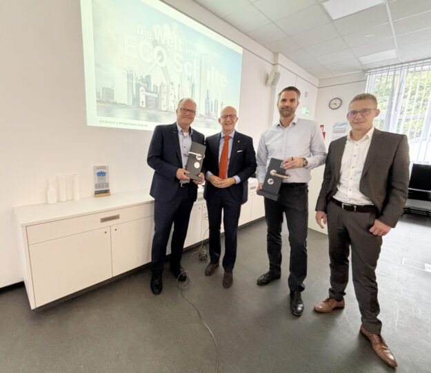 Vier Männer stehen in einem Besprechungsraum vor einer Präsentation mit der Aufschrift „Die Welt von ECO Schulte“. Zwei von ihnen halten schwarze Präsentationsboxen in den Händen.