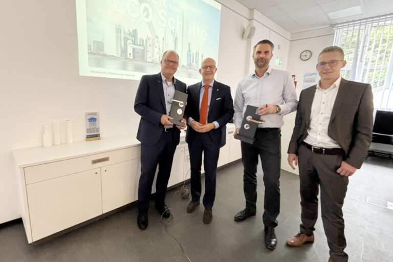 Vier Männer stehen in einem Besprechungsraum vor einer Präsentation mit der Aufschrift „Die Welt von ECO Schulte“. Zwei von ihnen halten schwarze Präsentationsboxen in den Händen.