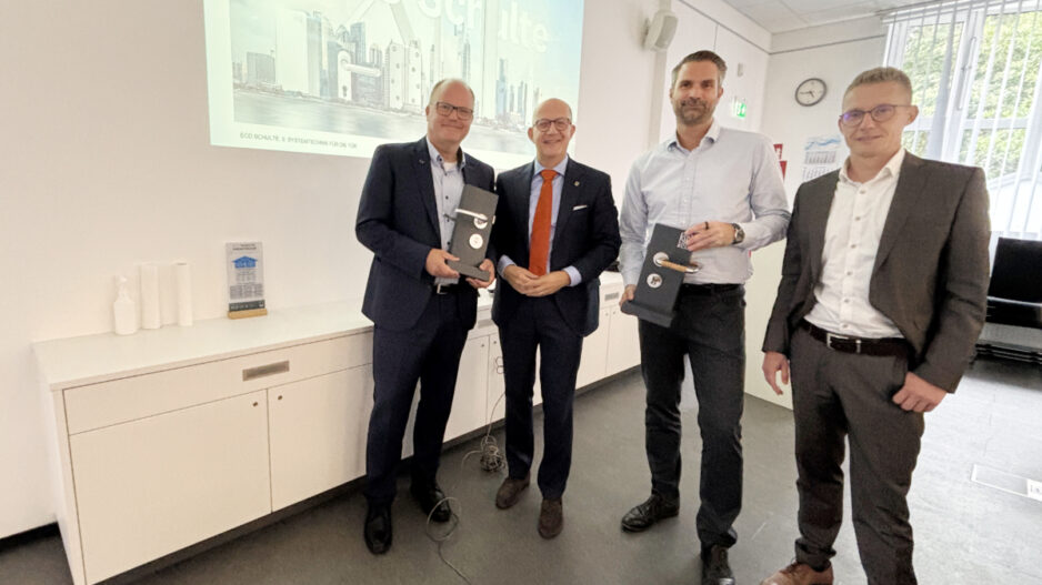Vier Männer stehen in einem Besprechungsraum vor einer Präsentation mit der Aufschrift „Die Welt von ECO Schulte“. Zwei von ihnen halten schwarze Präsentationsboxen in den Händen.