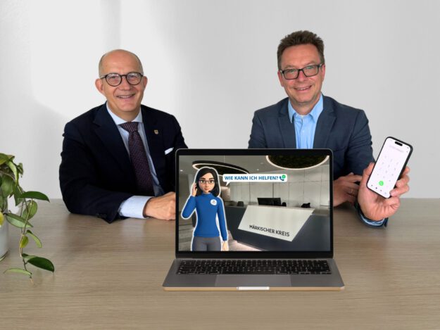 Zwei Männer im Anzug sitzen lächelnd an einem Tisch. Vor ihnen steht ein Laptop, auf dessen Bildschirm eine digitale Assistentin vor dem Empfang des Märkischen Kreises mit der Frage „Wie kann ich helfen?“ zu sehen ist. Einer der Männer hält ein Smartphone mit geöffneter Telefonfunktion in die Kamera.