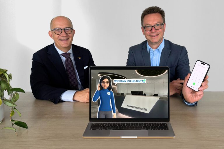 Zwei Männer im Anzug sitzen lächelnd an einem Tisch. Vor ihnen steht ein Laptop, auf dessen Bildschirm eine digitale Assistentin vor dem Empfang des Märkischen Kreises mit der Frage „Wie kann ich helfen?“ zu sehen ist. Einer der Männer hält ein Smartphone mit geöffneter Telefonfunktion in die Kamera.