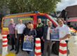 Eine Gruppe von sieben Personen steht vor einem roten Einsatzleitungsfahrzeug von Feuerwehr und Polizei. Zwei Personen halten Plakate zum „1. Mendener Blaulichttag“ am 19. September 2026 in der Mendener Innenstadt.