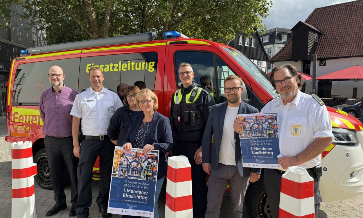 Eine Gruppe von sieben Personen steht vor einem roten Einsatzleitungsfahrzeug von Feuerwehr und Polizei. Zwei Personen halten Plakate zum „1. Mendener Blaulichttag“ am 19. September 2026 in der Mendener Innenstadt.