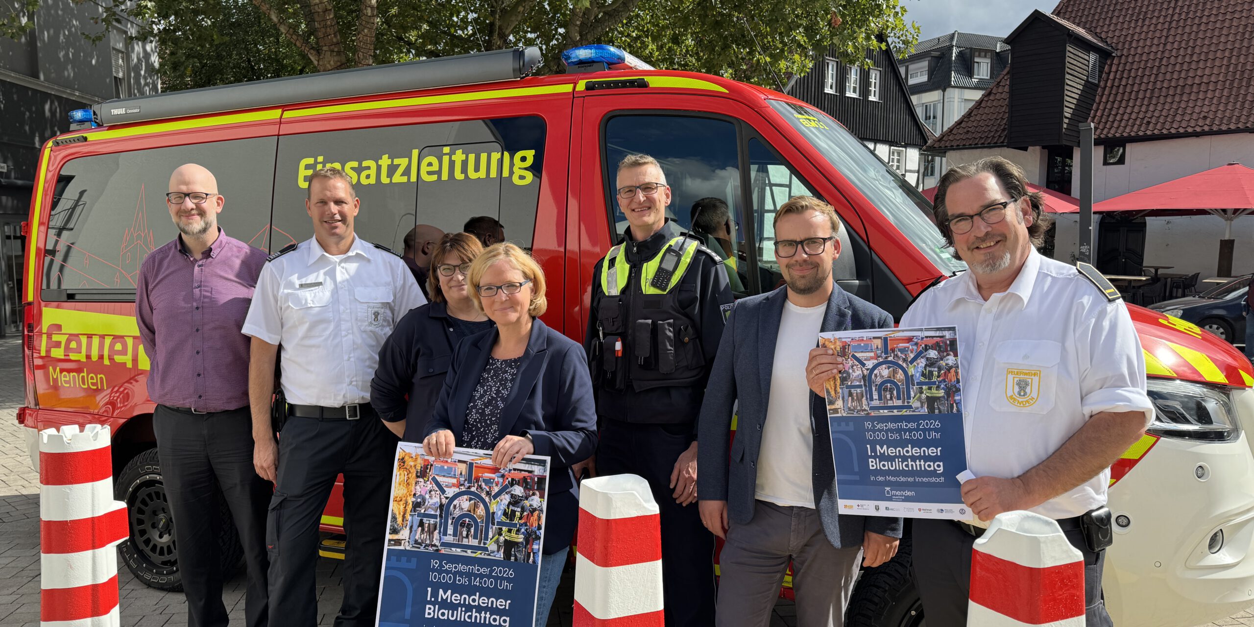 Eine Gruppe von sieben Personen steht vor einem roten Einsatzleitungsfahrzeug von Feuerwehr und Polizei. Zwei Personen halten Plakate zum „1. Mendener Blaulichttag“ am 19. September 2026 in der Mendener Innenstadt.