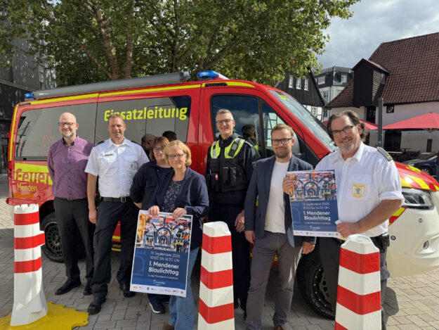 Eine Gruppe von sieben Personen steht vor einem roten Einsatzleitungsfahrzeug von Feuerwehr und Polizei. Zwei Personen halten Plakate zum „1. Mendener Blaulichttag“ am 19. September 2026 in der Mendener Innenstadt.