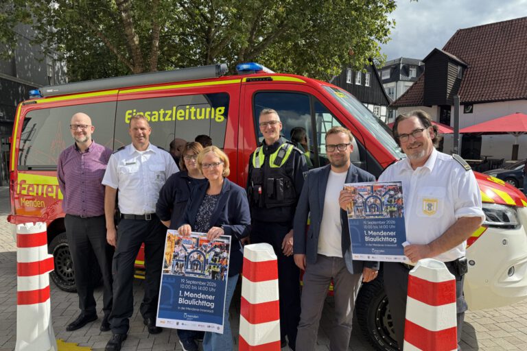 Eine Gruppe von sieben Personen steht vor einem roten Einsatzleitungsfahrzeug von Feuerwehr und Polizei. Zwei Personen halten Plakate zum „1. Mendener Blaulichttag“ am 19. September 2026 in der Mendener Innenstadt.