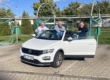 Drei Männer reinigen bei sonnigem Wetter gemeinsam ein weißes Cabriolet in einer Selbstbedienungs-Waschanlage. Sie stehen lachend am geöffneten Fahrzeug und putzen Scheiben und Karosserie.