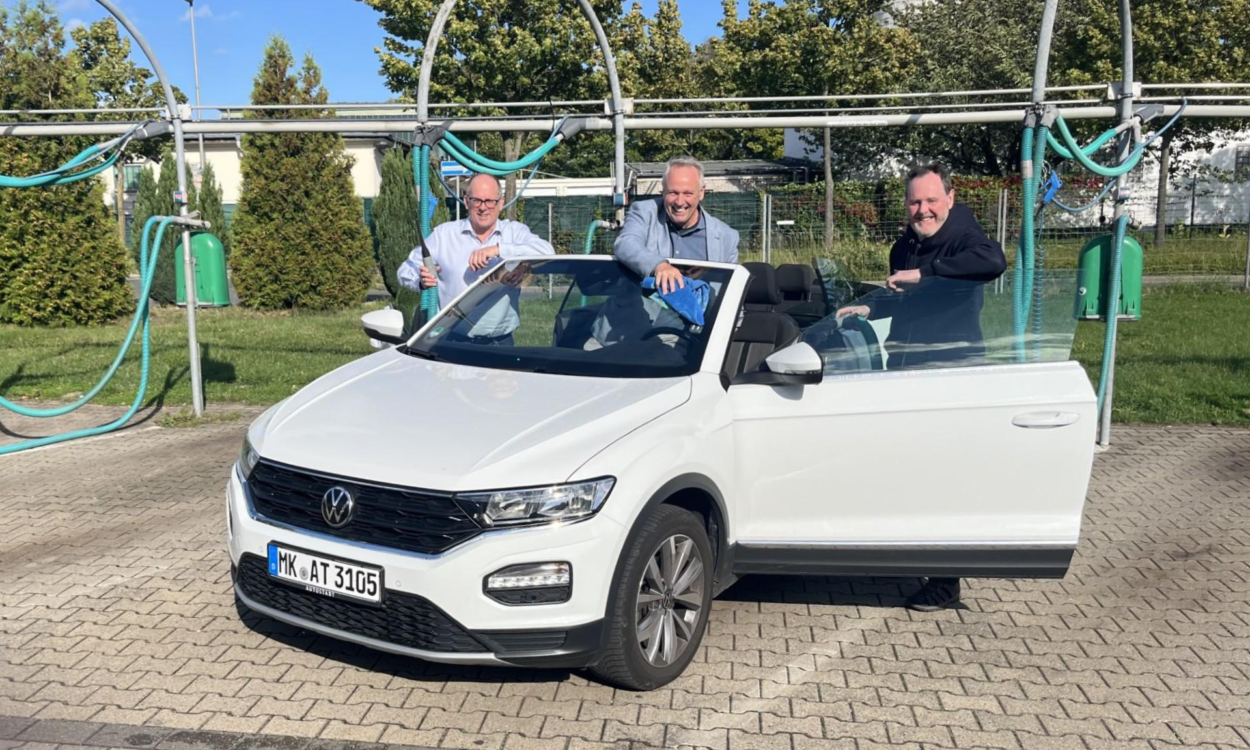 Drei Männer reinigen bei sonnigem Wetter gemeinsam ein weißes Cabriolet in einer Selbstbedienungs-Waschanlage. Sie stehen lachend am geöffneten Fahrzeug und putzen Scheiben und Karosserie.