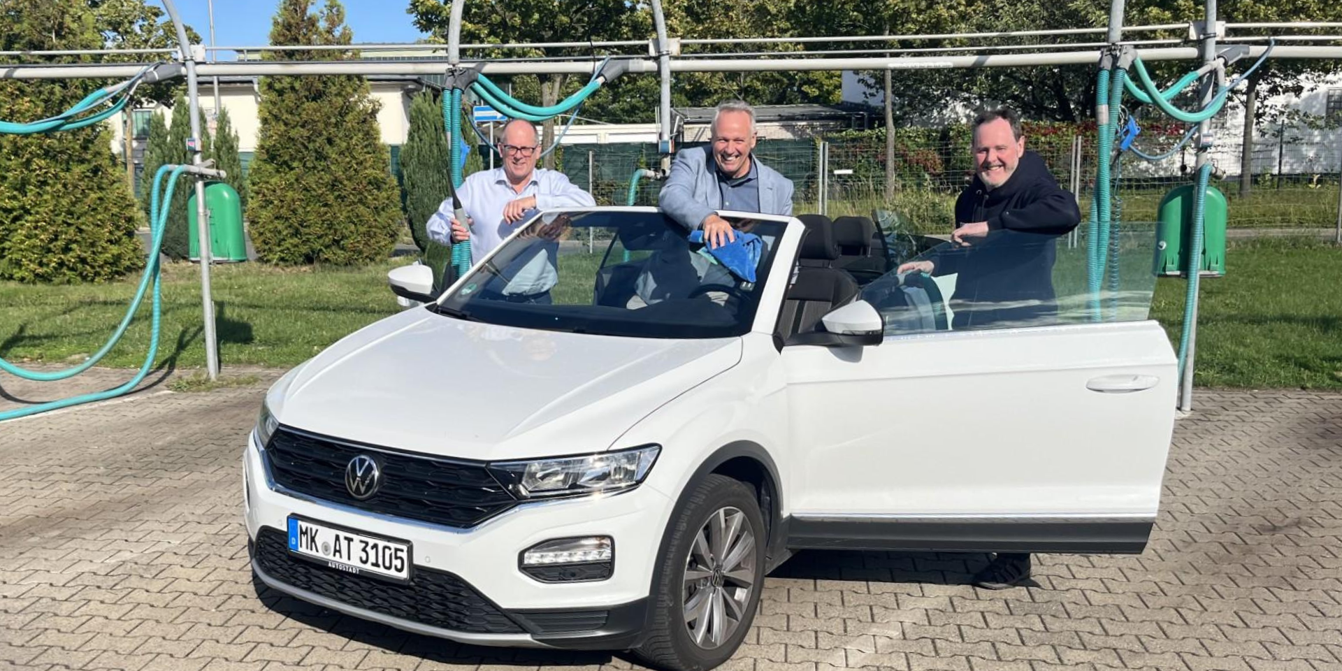 Drei Männer reinigen bei sonnigem Wetter gemeinsam ein weißes Cabriolet in einer Selbstbedienungs-Waschanlage. Sie stehen lachend am geöffneten Fahrzeug und putzen Scheiben und Karosserie.