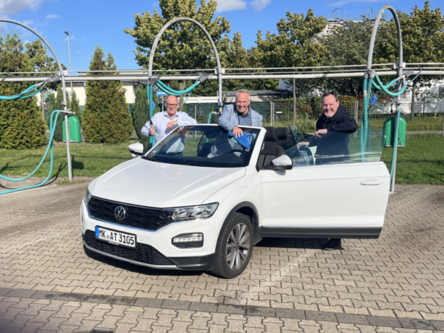 Drei Männer reinigen bei sonnigem Wetter gemeinsam ein weißes Cabriolet in einer Selbstbedienungs-Waschanlage. Sie stehen lachend am geöffneten Fahrzeug und putzen Scheiben und Karosserie.