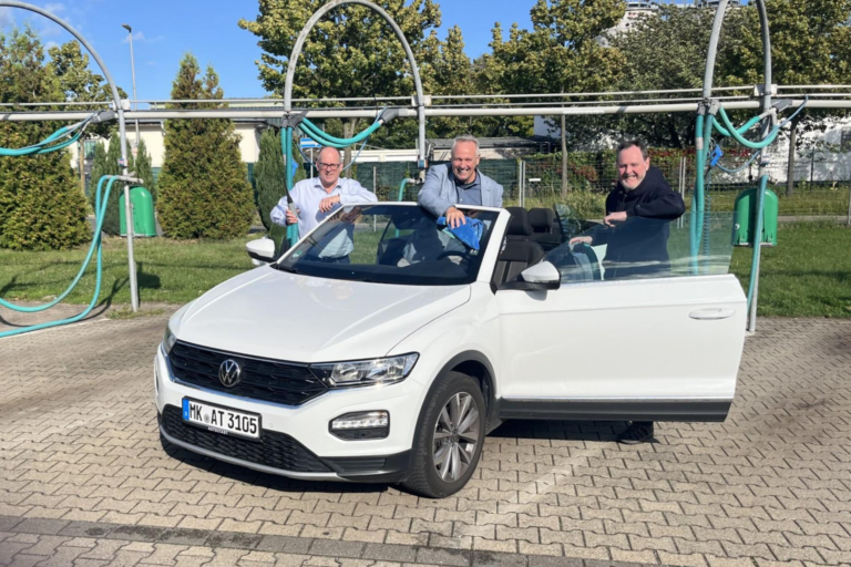 Drei Männer reinigen bei sonnigem Wetter gemeinsam ein weißes Cabriolet in einer Selbstbedienungs-Waschanlage. Sie stehen lachend am geöffneten Fahrzeug und putzen Scheiben und Karosserie.