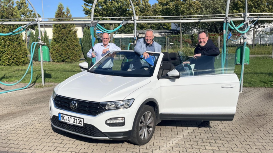Drei Männer reinigen bei sonnigem Wetter gemeinsam ein weißes Cabriolet in einer Selbstbedienungs-Waschanlage. Sie stehen lachend am geöffneten Fahrzeug und putzen Scheiben und Karosserie.