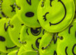 Nahaufnahme eines Stapels leuchtend gelber, runder Reflektoren mit verschiedenen aufgedruckten Smiley-Gesichtern. Die Anhänger sind mit kleinen Kugelketten versehen und liegen übereinander auf einer roten Unterlage.