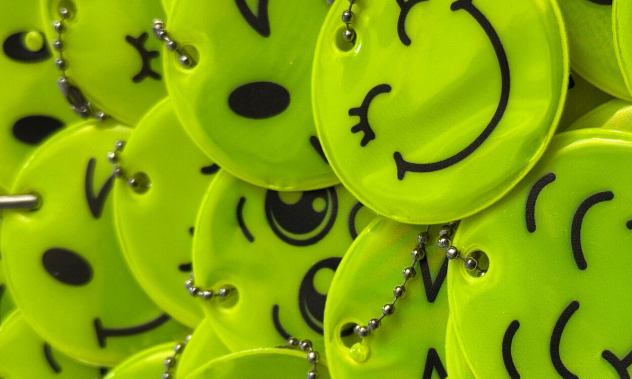 Nahaufnahme eines Stapels leuchtend gelber, runder Reflektoren mit verschiedenen aufgedruckten Smiley-Gesichtern. Die Anhänger sind mit kleinen Kugelketten versehen und liegen übereinander auf einer roten Unterlage.