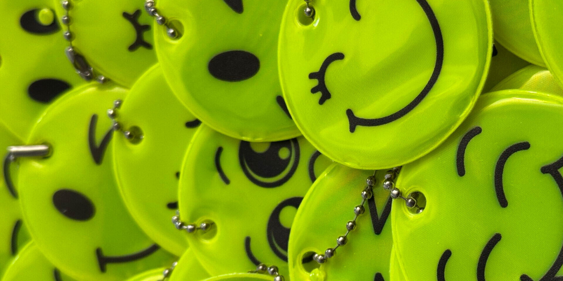 Nahaufnahme eines Stapels leuchtend gelber, runder Reflektoren mit verschiedenen aufgedruckten Smiley-Gesichtern. Die Anhänger sind mit kleinen Kugelketten versehen und liegen übereinander auf einer roten Unterlage.