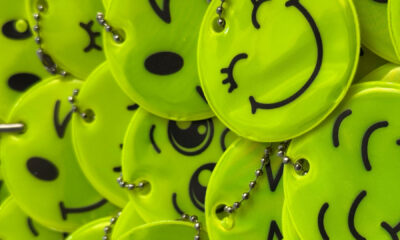 Nahaufnahme eines Stapels leuchtend gelber, runder Reflektoren mit verschiedenen aufgedruckten Smiley-Gesichtern. Die Anhänger sind mit kleinen Kugelketten versehen und liegen übereinander auf einer roten Unterlage.