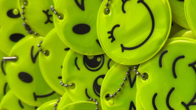 Nahaufnahme eines Stapels leuchtend gelber, runder Reflektoren mit verschiedenen aufgedruckten Smiley-Gesichtern. Die Anhänger sind mit kleinen Kugelketten versehen und liegen übereinander auf einer roten Unterlage.