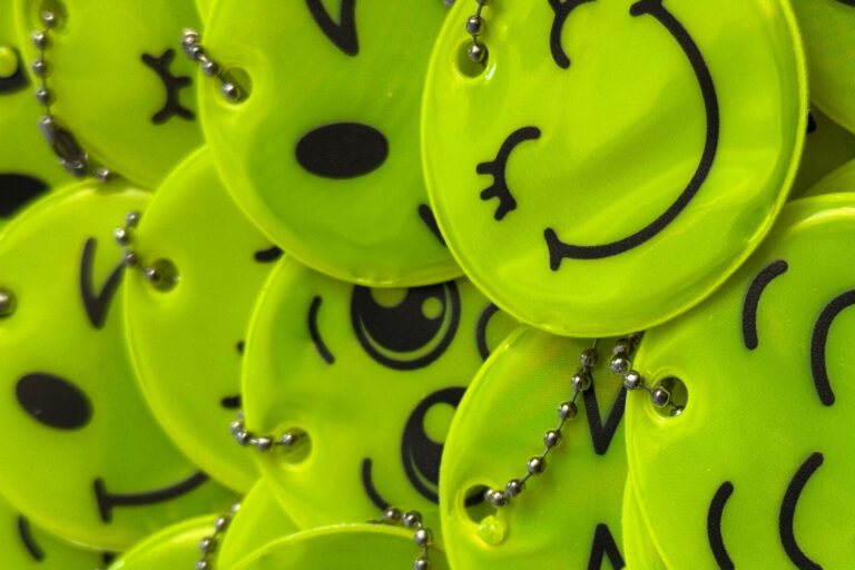 Nahaufnahme eines Stapels leuchtend gelber, runder Reflektoren mit verschiedenen aufgedruckten Smiley-Gesichtern. Die Anhänger sind mit kleinen Kugelketten versehen und liegen übereinander auf einer roten Unterlage.