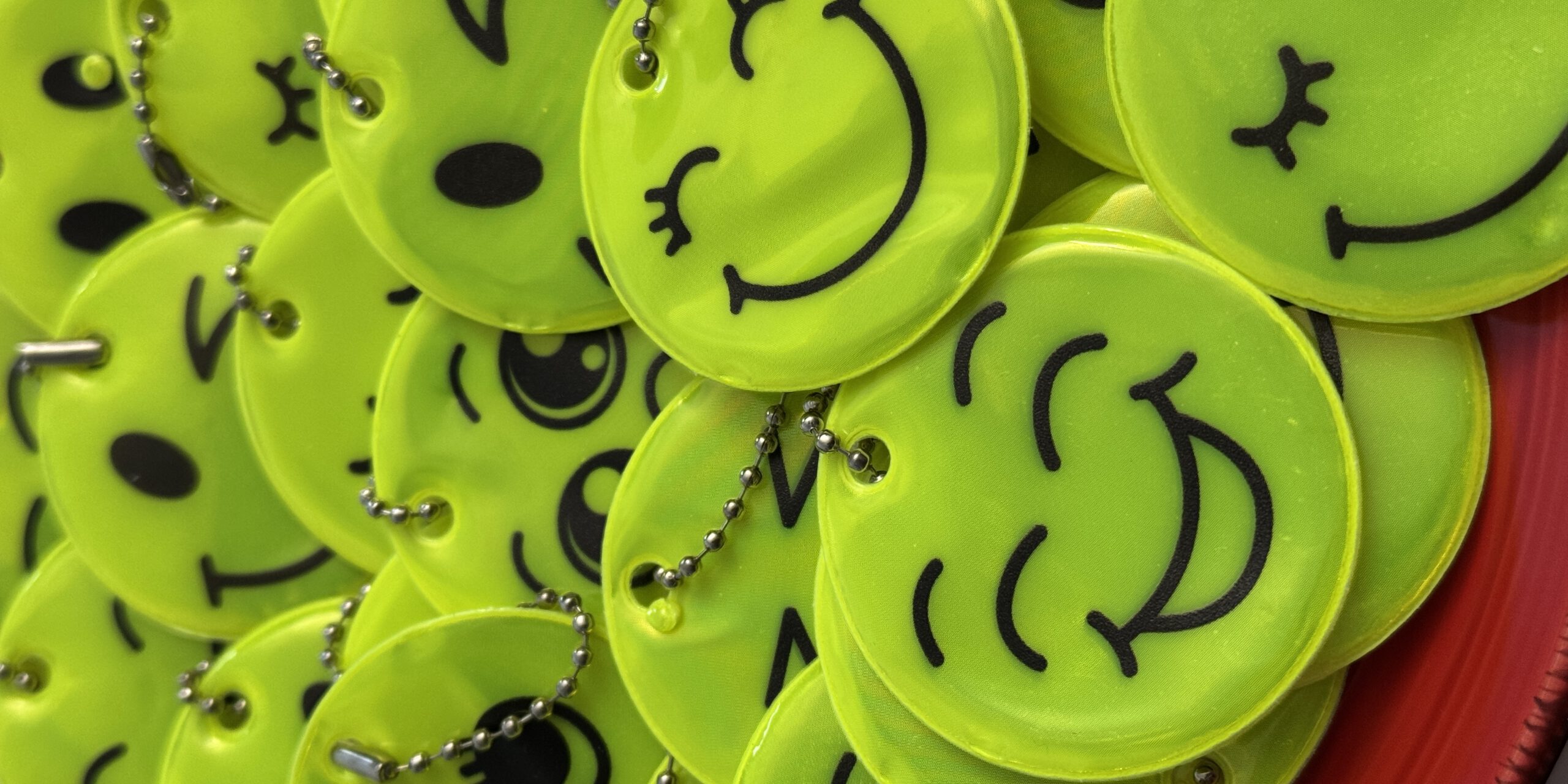Nahaufnahme eines Stapels leuchtend gelber, runder Reflektoren mit verschiedenen aufgedruckten Smiley-Gesichtern. Die Anhänger sind mit kleinen Kugelketten versehen und liegen übereinander auf einer roten Unterlage.