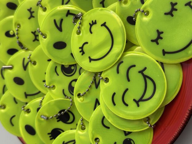 Nahaufnahme eines Stapels leuchtend gelber, runder Reflektoren mit verschiedenen aufgedruckten Smiley-Gesichtern. Die Anhänger sind mit kleinen Kugelketten versehen und liegen übereinander auf einer roten Unterlage.