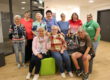 Gruppenfoto von zehn Frauen unterschiedlichen Alters in einem hellen Gemeinschaftsraum. Einige stehen, drei sitzen im Vordergrund. In der Mitte befindet sich eine große Kiste mit zahlreichen kleinen, bunt gestrickten Söckchen; mehrere Frauen halten einzelne Söckchen in die Kamera.
