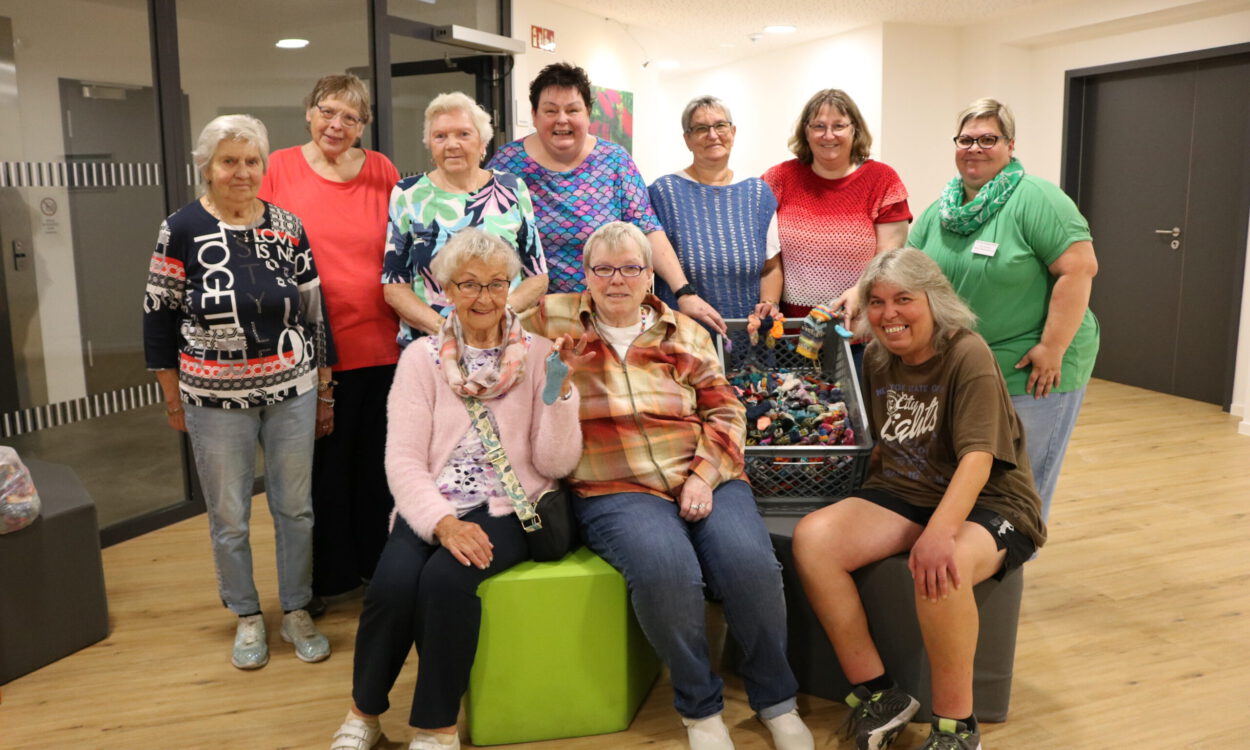 Gruppenfoto von zehn Frauen unterschiedlichen Alters in einem hellen Gemeinschaftsraum. Einige stehen, drei sitzen im Vordergrund. In der Mitte befindet sich eine große Kiste mit zahlreichen kleinen, bunt gestrickten Söckchen; mehrere Frauen halten einzelne Söckchen in die Kamera.