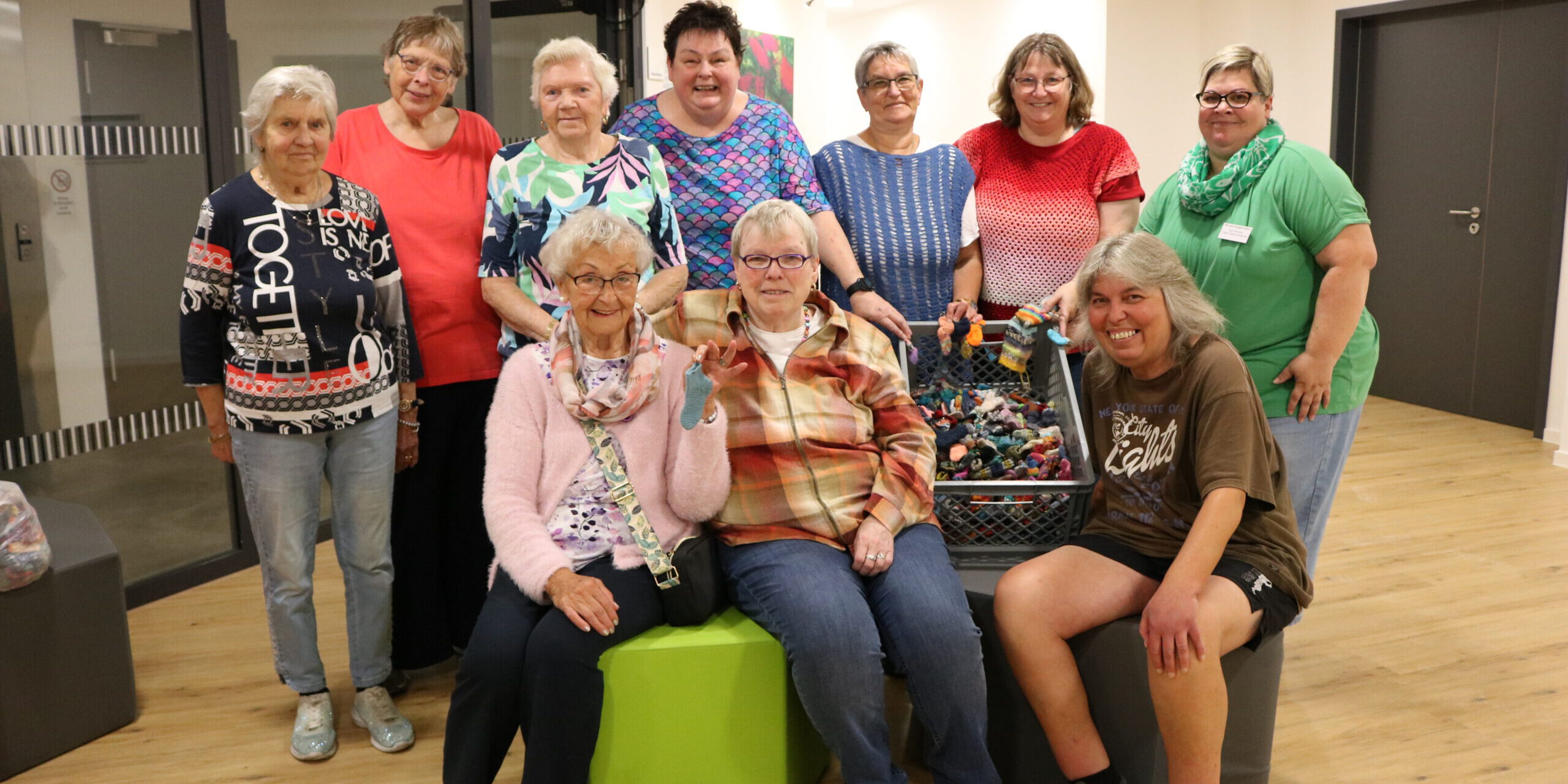 Gruppenfoto von zehn Frauen unterschiedlichen Alters in einem hellen Gemeinschaftsraum. Einige stehen, drei sitzen im Vordergrund. In der Mitte befindet sich eine große Kiste mit zahlreichen kleinen, bunt gestrickten Söckchen; mehrere Frauen halten einzelne Söckchen in die Kamera.