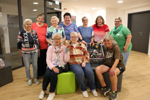 Gruppenfoto von zehn Frauen unterschiedlichen Alters in einem hellen Gemeinschaftsraum. Einige stehen, drei sitzen im Vordergrund. In der Mitte befindet sich eine große Kiste mit zahlreichen kleinen, bunt gestrickten Söckchen; mehrere Frauen halten einzelne Söckchen in die Kamera.