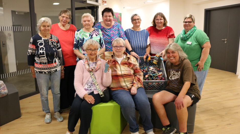 Gruppenfoto von zehn Frauen unterschiedlichen Alters in einem hellen Gemeinschaftsraum. Einige stehen, drei sitzen im Vordergrund. In der Mitte befindet sich eine große Kiste mit zahlreichen kleinen, bunt gestrickten Söckchen; mehrere Frauen halten einzelne Söckchen in die Kamera.