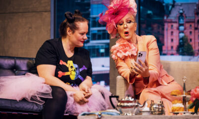 Zwei Frauen sitzen auf einer Theaterbühne in einer Wohnzimmerkulisse. Eine trägt einen auffälligen rosafarbenen Hosenanzug mit großem Blumenhut und zeigt der anderen etwas auf ihrem Smartphone; vor ihnen steht ein Tisch mit Teegeschirr und Gebäck.