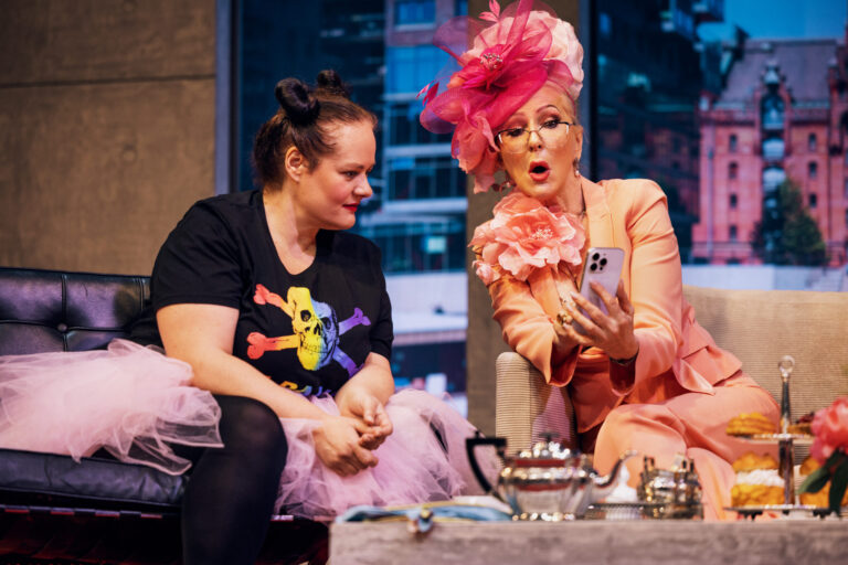 Zwei Frauen sitzen auf einer Theaterbühne in einer Wohnzimmerkulisse. Eine trägt einen auffälligen rosafarbenen Hosenanzug mit großem Blumenhut und zeigt der anderen etwas auf ihrem Smartphone; vor ihnen steht ein Tisch mit Teegeschirr und Gebäck.