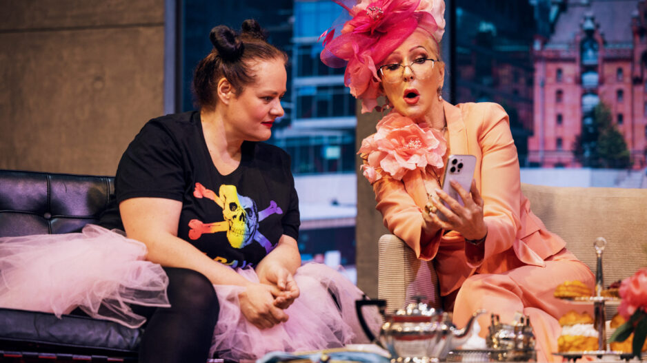 Zwei Frauen sitzen auf einer Theaterbühne in einer Wohnzimmerkulisse. Eine trägt einen auffälligen rosafarbenen Hosenanzug mit großem Blumenhut und zeigt der anderen etwas auf ihrem Smartphone; vor ihnen steht ein Tisch mit Teegeschirr und Gebäck.