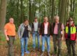 Sechs Männer stehen nebeneinander auf einem mit Laub bedeckten Waldboden. Sie tragen überwiegend Freizeit- und Forstkleidung; im Hintergrund sind zahlreiche hohe Laubbäume zu sehen.