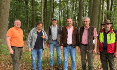 Sechs Männer stehen nebeneinander auf einem mit Laub bedeckten Waldboden. Sie tragen überwiegend Freizeit- und Forstkleidung; im Hintergrund sind zahlreiche hohe Laubbäume zu sehen.