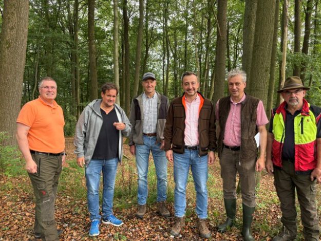 Sechs Männer stehen nebeneinander auf einem mit Laub bedeckten Waldboden. Sie tragen überwiegend Freizeit- und Forstkleidung; im Hintergrund sind zahlreiche hohe Laubbäume zu sehen.