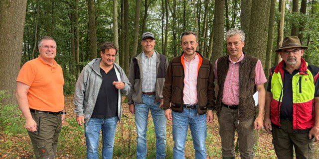 Sechs Männer stehen nebeneinander auf einem mit Laub bedeckten Waldboden. Sie tragen überwiegend Freizeit- und Forstkleidung; im Hintergrund sind zahlreiche hohe Laubbäume zu sehen.