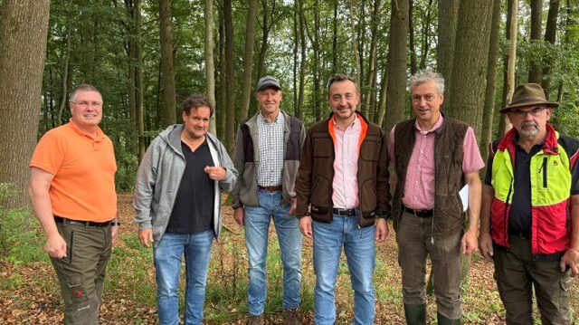 Sechs Männer stehen nebeneinander auf einem mit Laub bedeckten Waldboden. Sie tragen überwiegend Freizeit- und Forstkleidung; im Hintergrund sind zahlreiche hohe Laubbäume zu sehen.