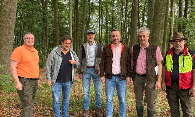 Sechs Männer stehen nebeneinander auf einem mit Laub bedeckten Waldboden. Sie tragen überwiegend Freizeit- und Forstkleidung; im Hintergrund sind zahlreiche hohe Laubbäume zu sehen.