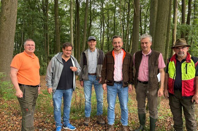 Sechs Männer stehen nebeneinander auf einem mit Laub bedeckten Waldboden. Sie tragen überwiegend Freizeit- und Forstkleidung; im Hintergrund sind zahlreiche hohe Laubbäume zu sehen.