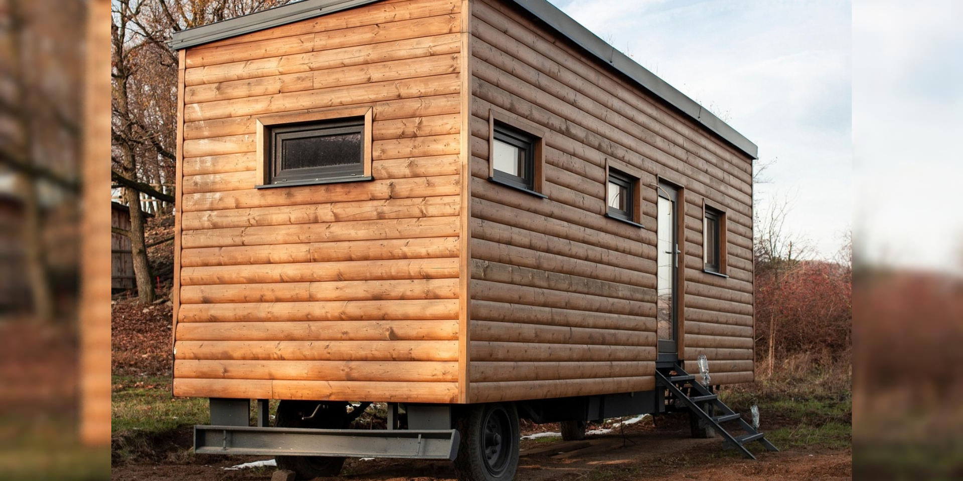Ein mobiles Tiny House mit Holzfassade steht auf einem Fahrgestell in einer naturnahen Umgebung. Eine kleine Treppe führt zur seitlichen Eingangstür; mehrere Fenster sind in die Holzfassade eingelassen.
