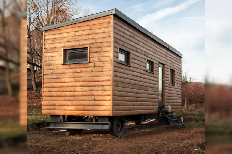 Ein mobiles Tiny House mit Holzfassade steht auf einem Fahrgestell in einer naturnahen Umgebung. Eine kleine Treppe führt zur seitlichen Eingangstür; mehrere Fenster sind in die Holzfassade eingelassen.