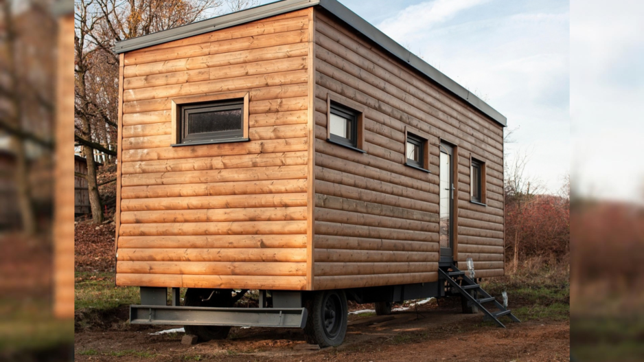 Ein mobiles Tiny House mit Holzfassade steht auf einem Fahrgestell in einer naturnahen Umgebung. Eine kleine Treppe führt zur seitlichen Eingangstür; mehrere Fenster sind in die Holzfassade eingelassen.