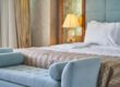 Modern eingerichtetes Hotelzimmer mit einem großen, frisch bezogenen Bett, weißen Kissen und einer gemusterten Tagesdecke. Am Fußende steht eine hellblaue Polsterbank; im Hintergrund sorgt eine eingeschaltete Nachttischlampe für warmes Licht.