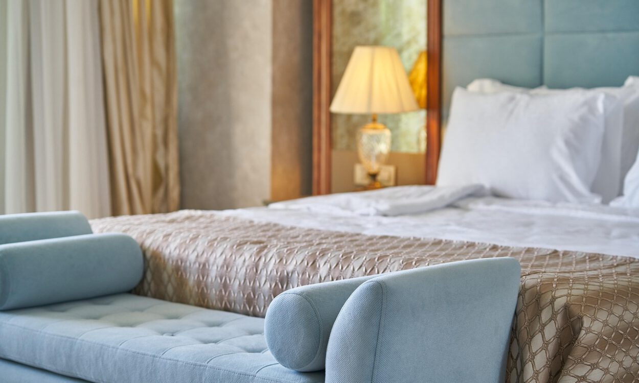 Modern eingerichtetes Hotelzimmer mit einem großen, frisch bezogenen Bett, weißen Kissen und einer gemusterten Tagesdecke. Am Fußende steht eine hellblaue Polsterbank; im Hintergrund sorgt eine eingeschaltete Nachttischlampe für warmes Licht.