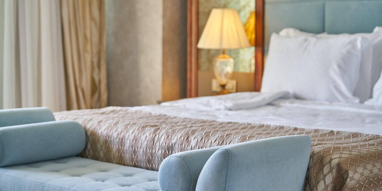 Modern eingerichtetes Hotelzimmer mit einem großen, frisch bezogenen Bett, weißen Kissen und einer gemusterten Tagesdecke. Am Fußende steht eine hellblaue Polsterbank; im Hintergrund sorgt eine eingeschaltete Nachttischlampe für warmes Licht.