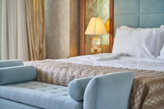 Modern eingerichtetes Hotelzimmer mit einem großen, frisch bezogenen Bett, weißen Kissen und einer gemusterten Tagesdecke. Am Fußende steht eine hellblaue Polsterbank; im Hintergrund sorgt eine eingeschaltete Nachttischlampe für warmes Licht.