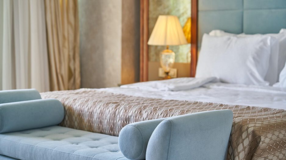Modern eingerichtetes Hotelzimmer mit einem großen, frisch bezogenen Bett, weißen Kissen und einer gemusterten Tagesdecke. Am Fußende steht eine hellblaue Polsterbank; im Hintergrund sorgt eine eingeschaltete Nachttischlampe für warmes Licht.