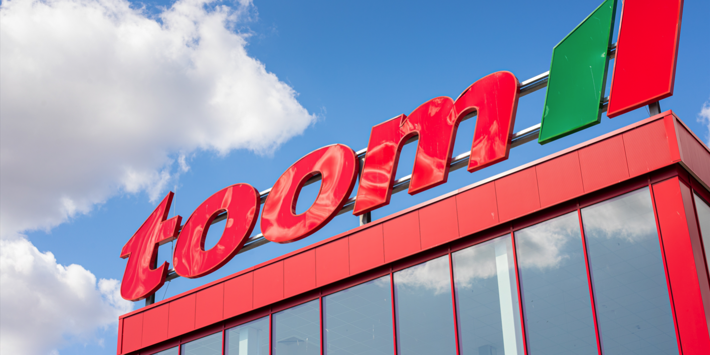Außenaufnahme eines toom-Baumarkts aus niedriger Perspektive. Über der rot verkleideten Glasfassade steht der große rote „toom“-Schriftzug mit grün-rotem Logo vor blauem Himmel und weißen Wolken.