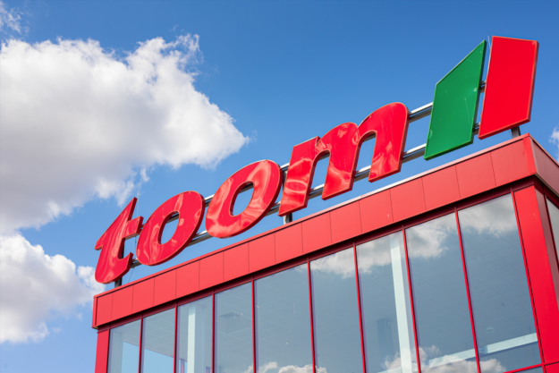 Außenaufnahme eines toom-Baumarkts aus niedriger Perspektive. Über der rot verkleideten Glasfassade steht der große rote „toom“-Schriftzug mit grün-rotem Logo vor blauem Himmel und weißen Wolken.